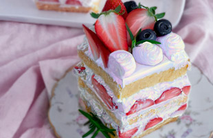 A digital image depicting a square slice of a strawberry and cream vanilla cake sitting on a floral plate. The cream has been colored with pastel colors. The background is a light pink cloth.
